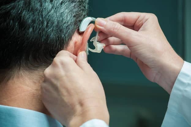 Close up of a hearing care specialist placing a behind-the-ear hearing aid on a man’s right ear. 
